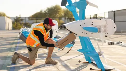 Amazon A worker kneels next to an Amazon drone and loads a package into its cargo bay. The drone is large - as big as the worker.