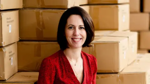 Irene Breen Irene Breen is pictured smiling. She has a dark brown bob haircut and is wearing a red shirt. She's sitting against stacks of boxes in a warehouse