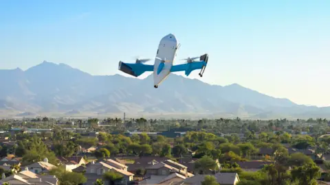 Amazon An Amazon drone flies over an US city. In the background are mountains and a blue sky.